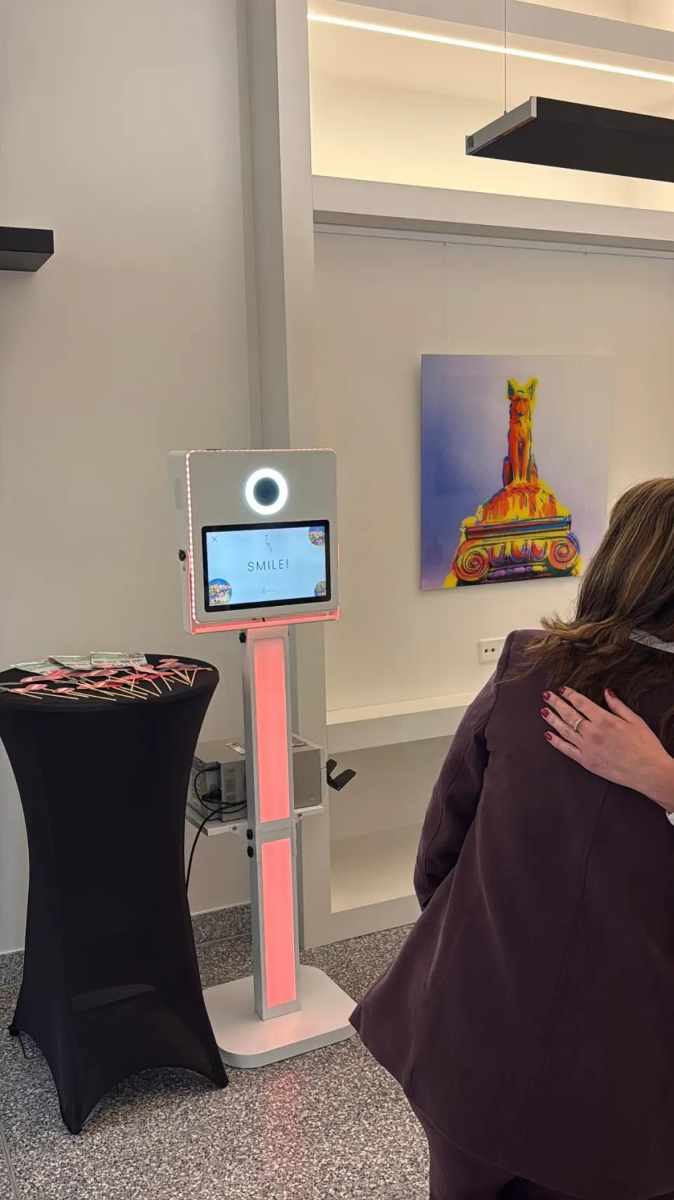 Borne photobooth lumineuse entière, colonne LED rose et écran « Smile! », accessoires sur un mange-debout, une invitée au premier plan