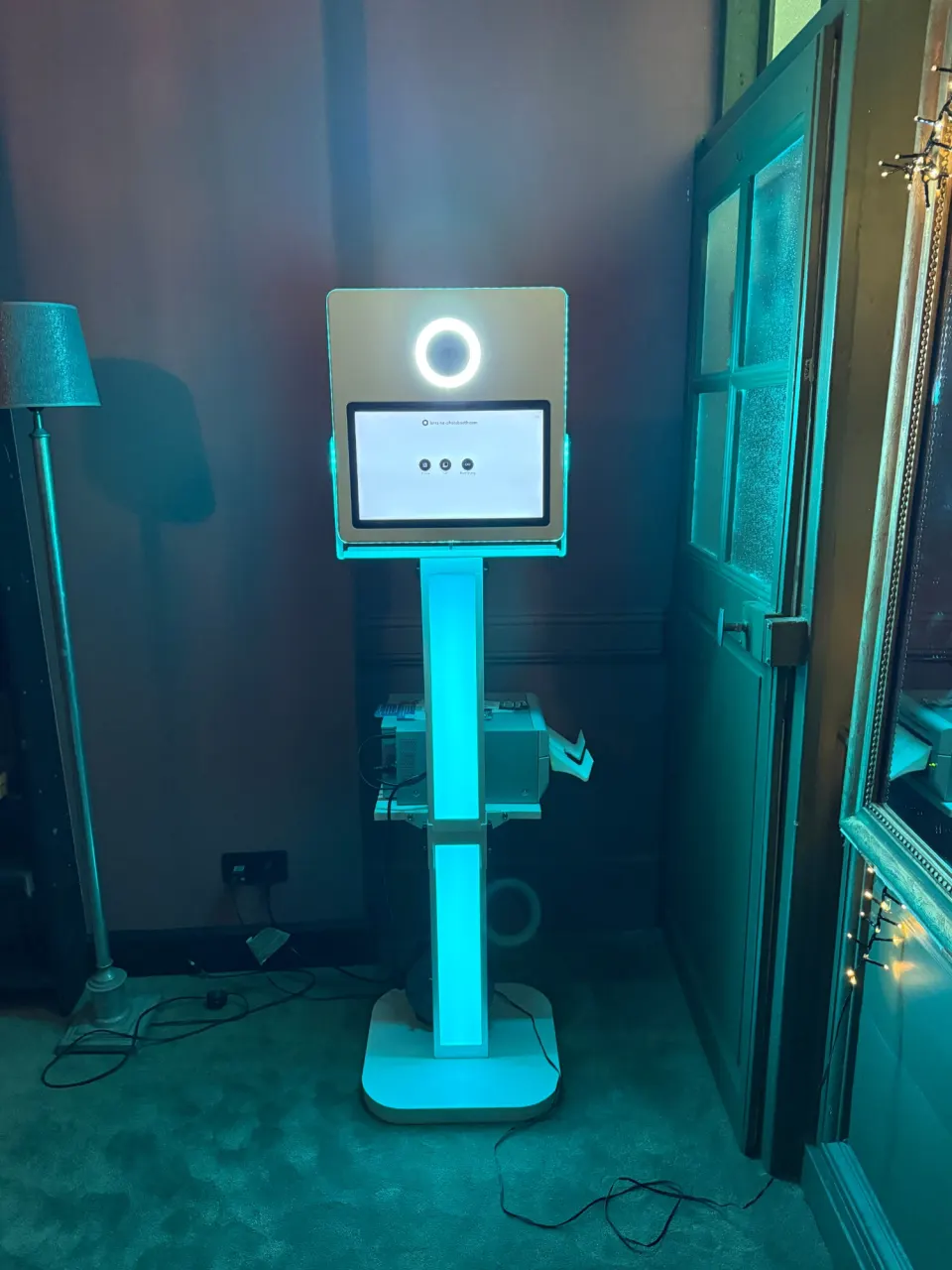Borne photobooth lumineuse entière, colonne LED turquoise et imprimante sous l'écran, dans une salle en soirée