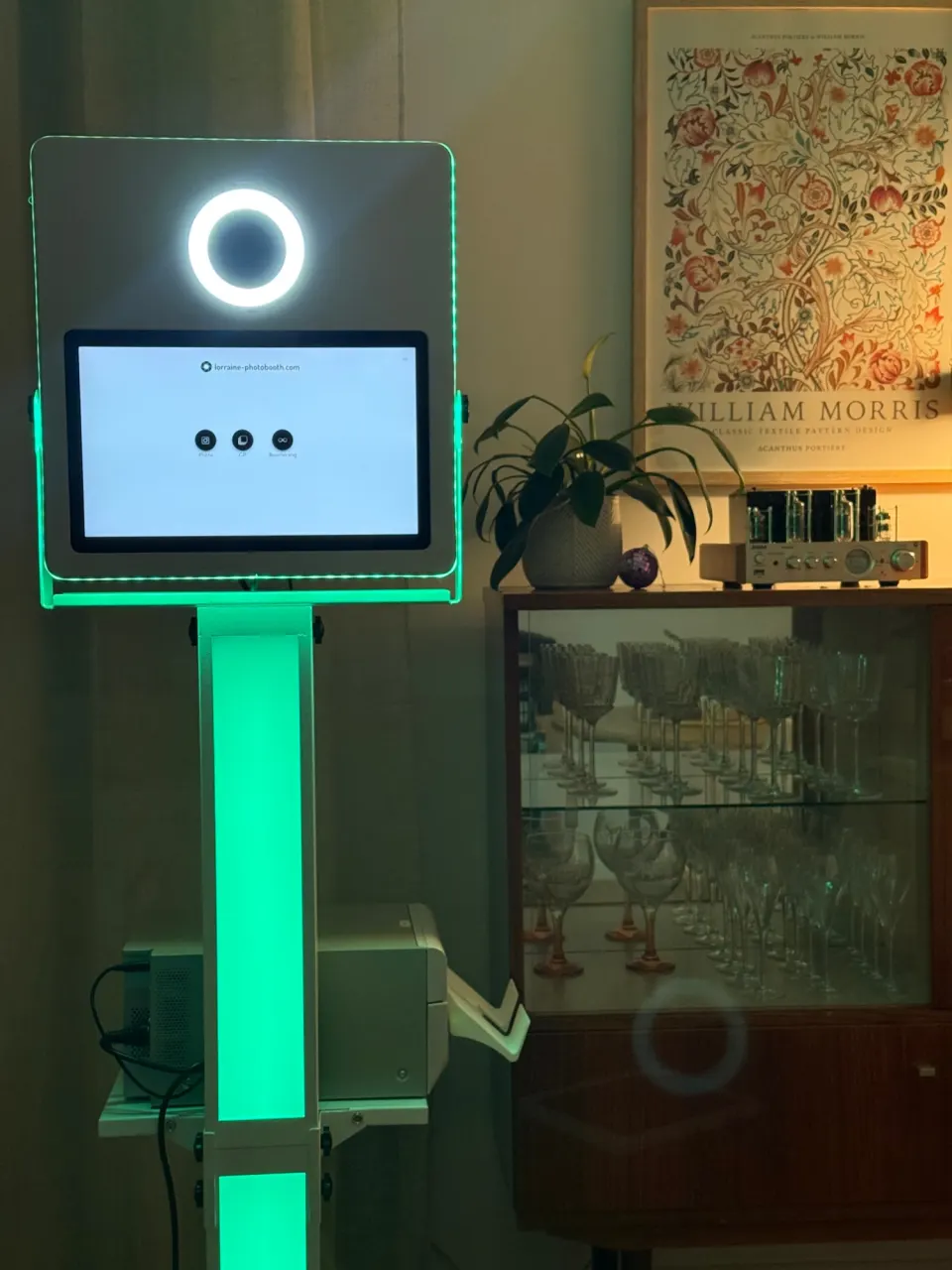 Borne photobooth lumineuse dans un salon, colonne LED verte, écran d'accueil et imprimante sous l'écran