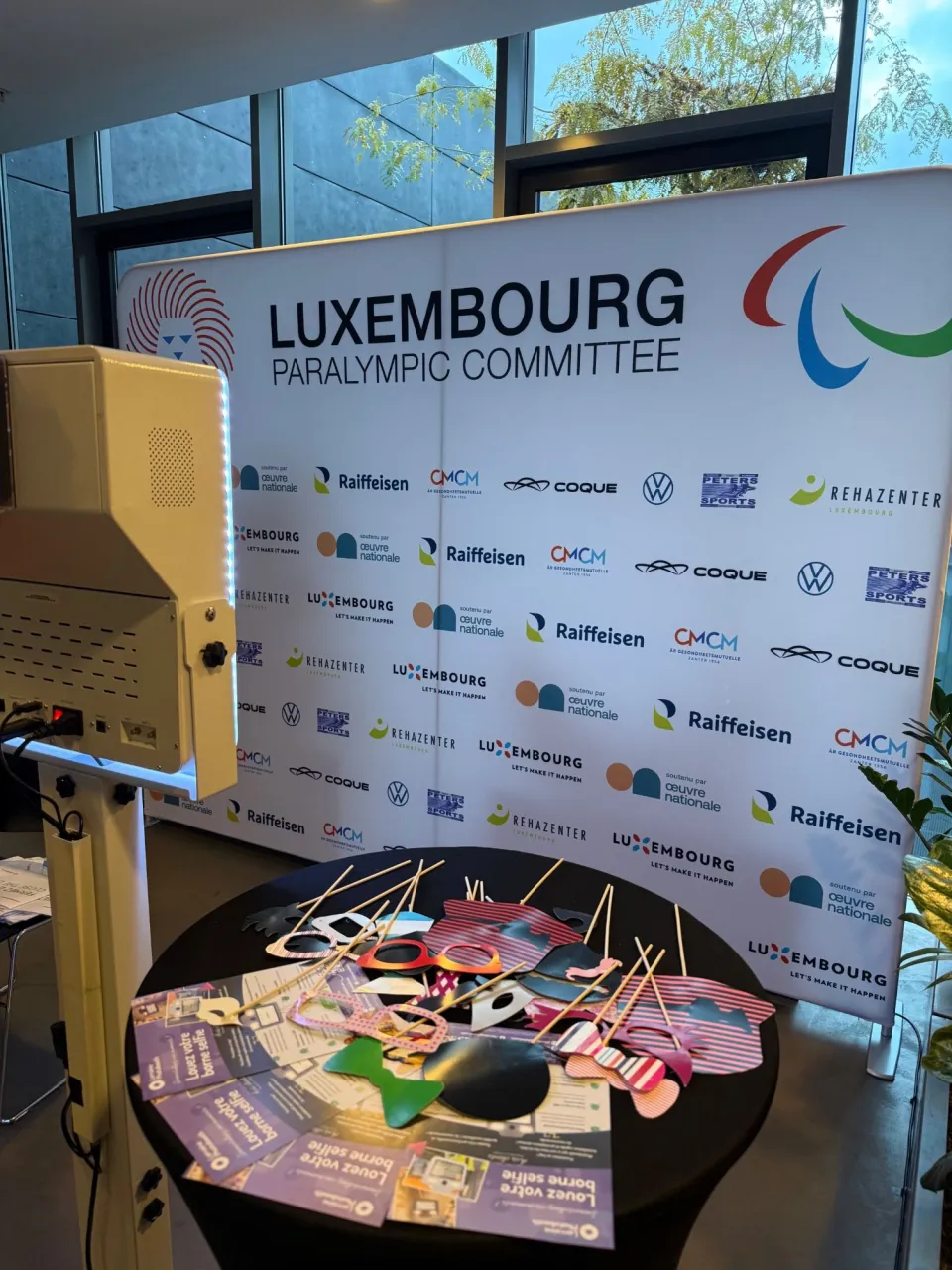 La borne vue de dos face au mur du Luxembourg Paralympic Committee et de ses partenaires, un mange-debout garni d'accessoires devant