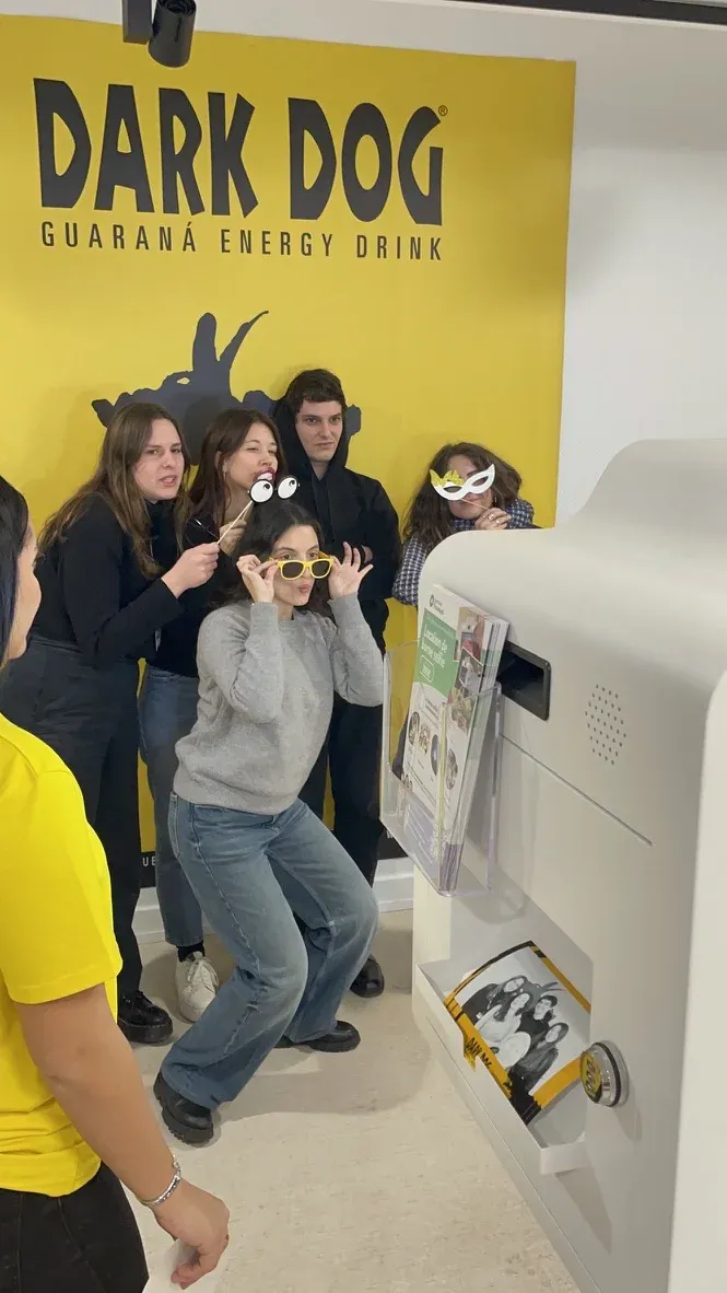 Un groupe pose avec lunettes et masques devant le mur d'une marque, le tirage précédent dans le bac de la borne